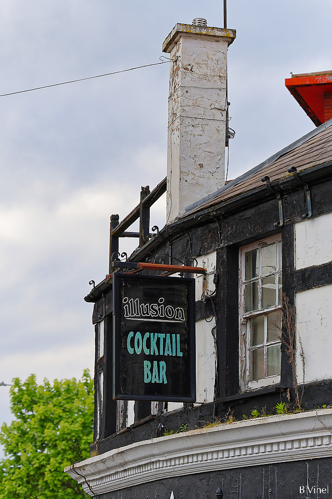 Illusion cocktail bar sign hanging from a half-timbered wall with a white chimney emerging from the roof