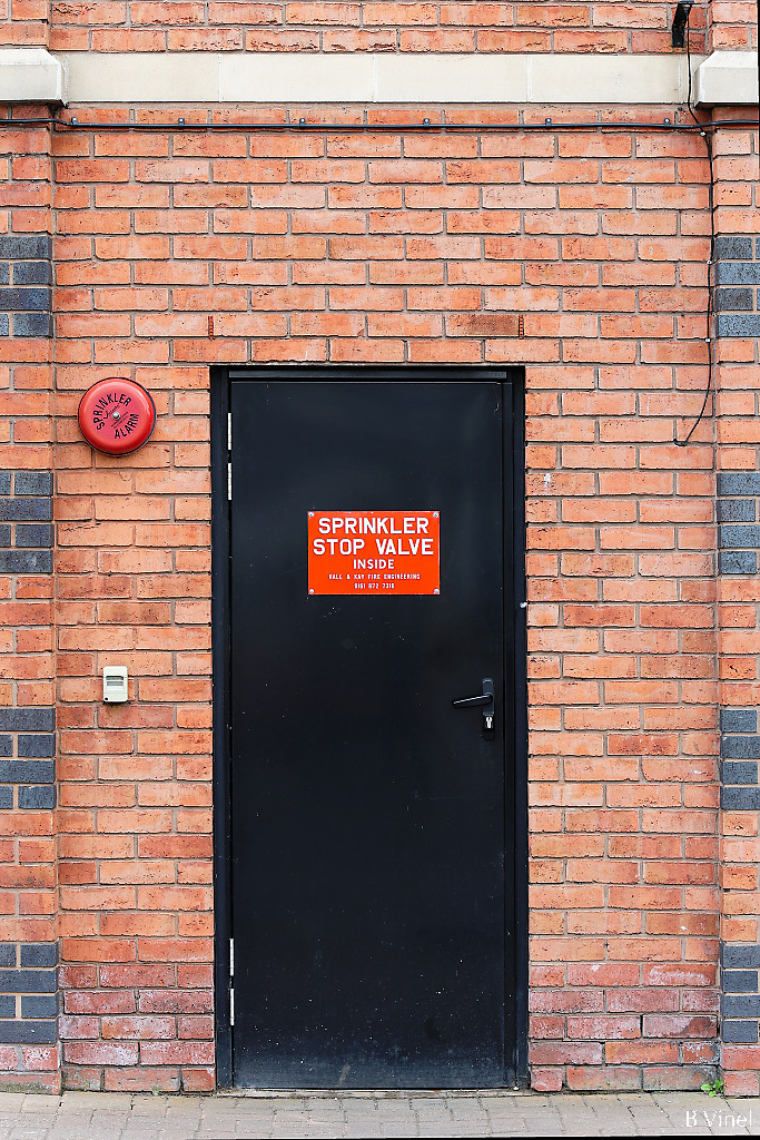 A black emergency door on a red brick wall with a red sign Sprinkle Stop Valve, and a red alarm bell on the side.