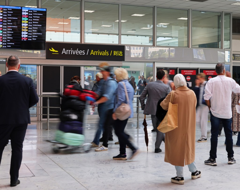 The picture shows the Nice Airport terminal 1 arrivals with people waiting for the passengers, including a gentleman with a suit and an umbrella, and few people leaving the terminal. They are blurred to illustrate that they are in movement.