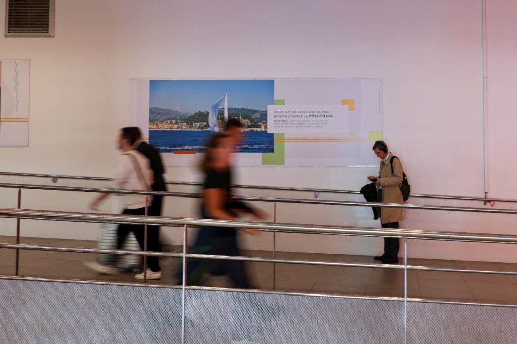 The picture was shot in the Nice Airport terminal 1 and show people departing and leaving the terminal, walking on a ramp. They are blurred to illustrate that they are in movement. There is a man standing still looking at his mobile phone.
