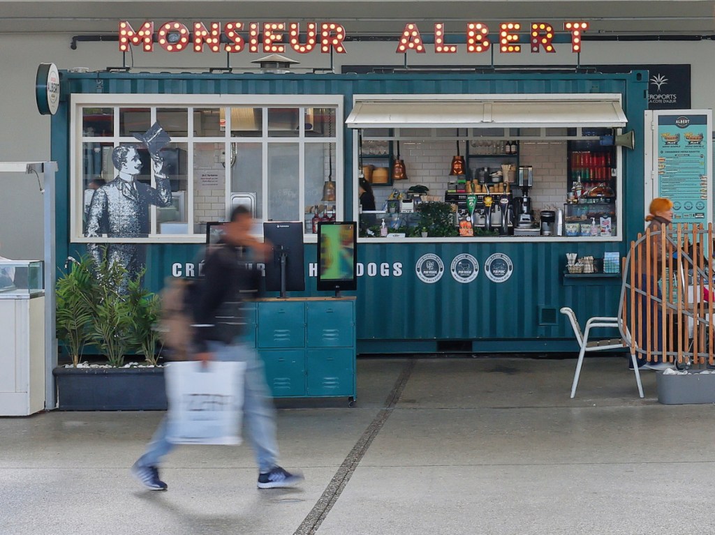 Nice Airport terminal 1. October 2024. The picture shows a blurred traveller with a white plastic bag and backpack, walking in front of a food stand called Monsieur Albert.