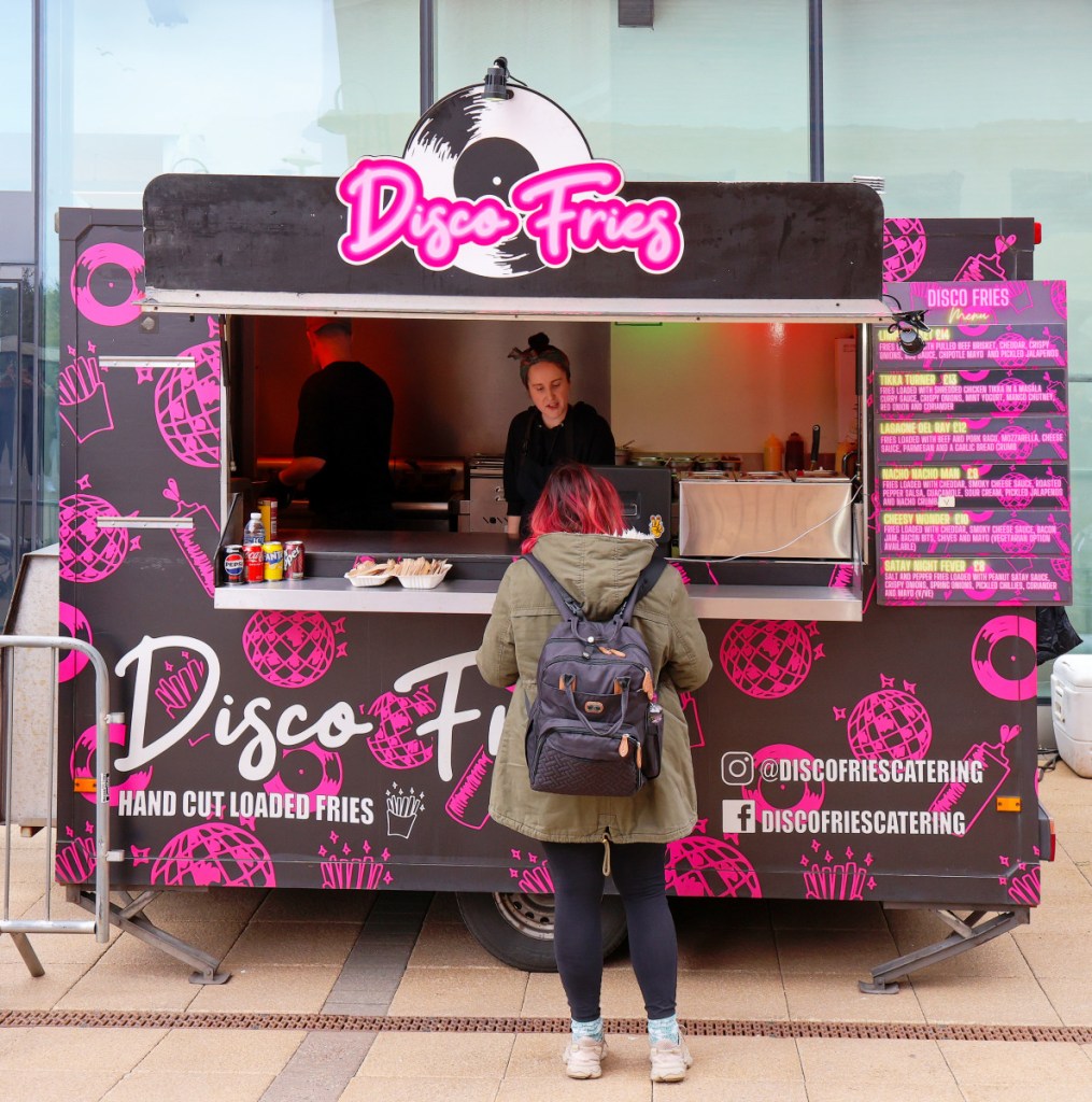 Image of trader in a fast food van branded Disco Fries and a customer in Kidderminster. The van is decorated with pink dancing balls and fries cones. There is a menu displayed on the side. The customer has red highlights in her hair and is carrying a backpack.