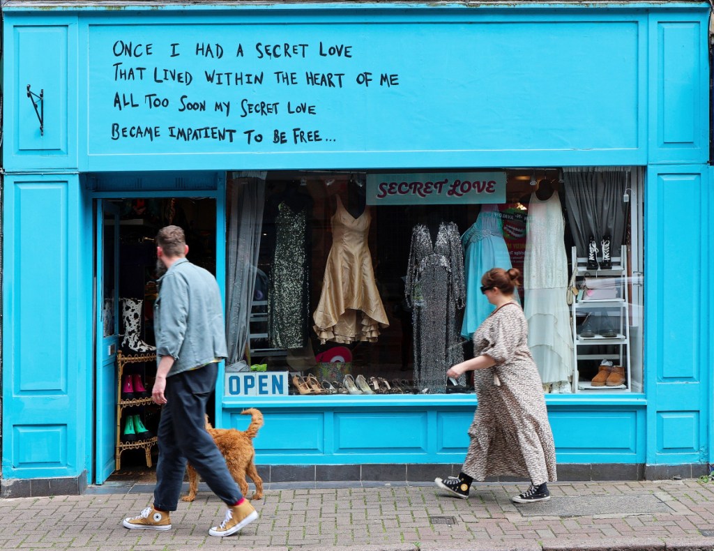 A man with a dog and a woman are walking in front of a thrift shop called Secret Love in Stourbridge. The shop front is painted light bright blue and there is quote handwritten on the fascia: "Once I had a script Love that lived within the heart of me. All too soon my secret love became impatient to be free". The window is well presented with elegant pre-loved women dresses and shoes.