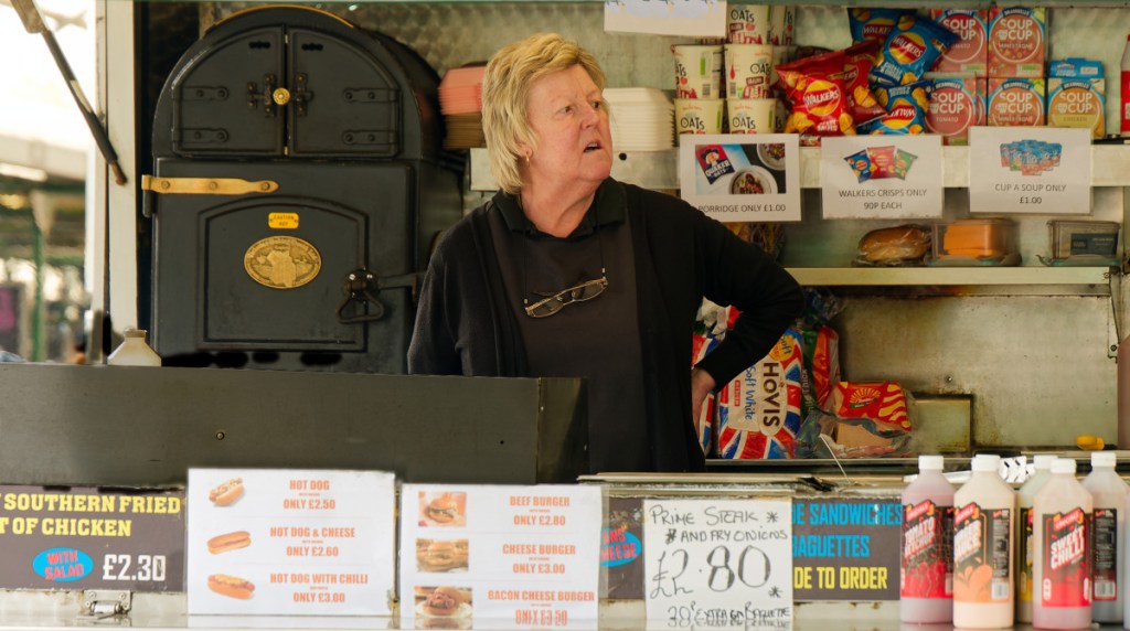 In a fast food van, a woman in her late sixties with blond hair is looking away. She is wearing black with a black apron, and her reading glasses hanging around her neck. There is a jacket potatoe oven behind her and multiple signs with food prices.