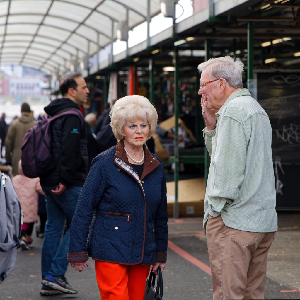 Customers in one of the aisles of the Bullring fruits and vegetables market. At the forefront, a woman with blond hair in the 70's style wears red trousers and a navy padded jacket, with colourful makeup. Next to her there is a man touching his mouth with his hand as if he was confused. Behind there is another man looking at the market stalls.