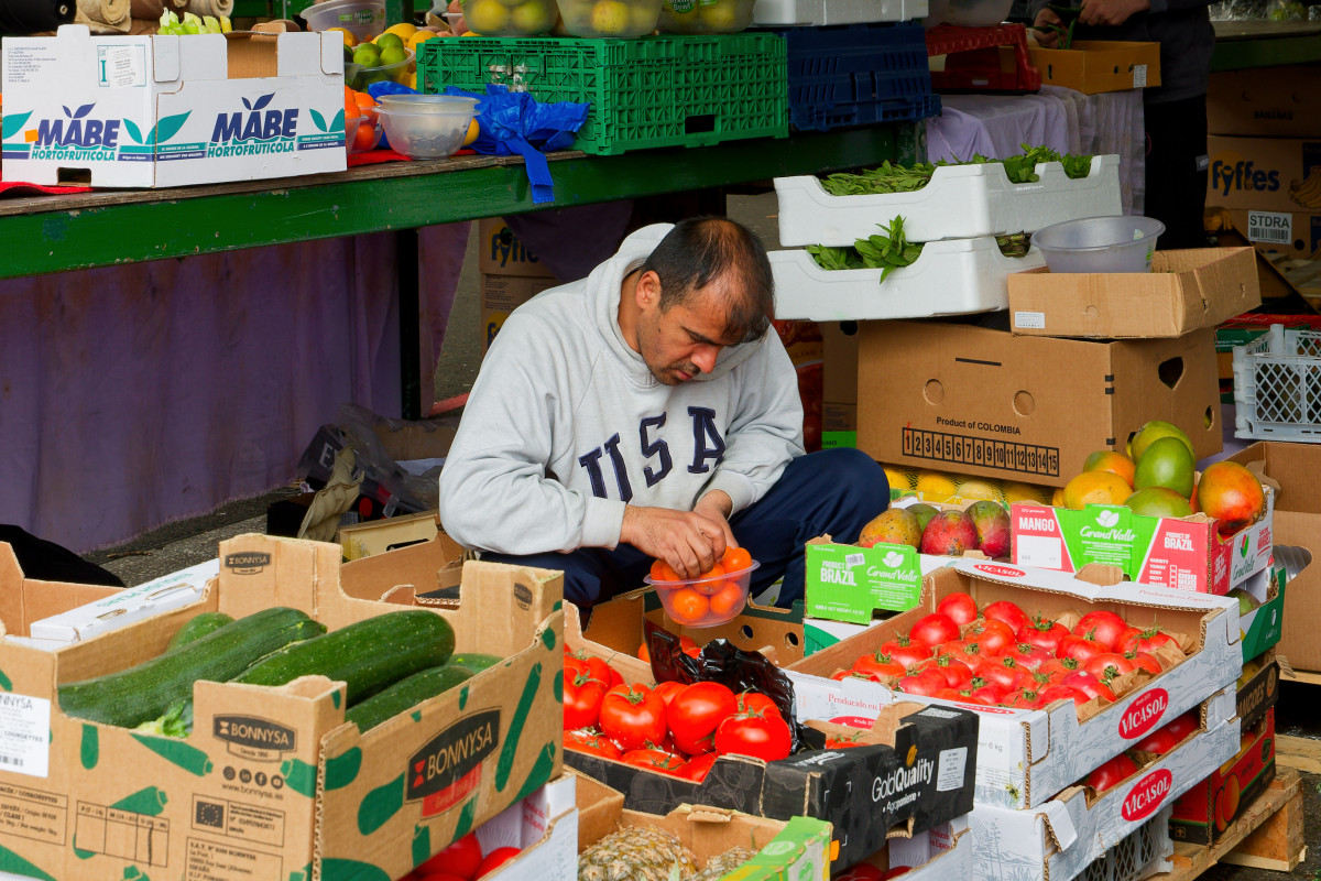 A asian man is crouching and packing fruits in a plastic bowl for sale at the Bullring Market. He is surrounded by boxes of fruits and vegetables.