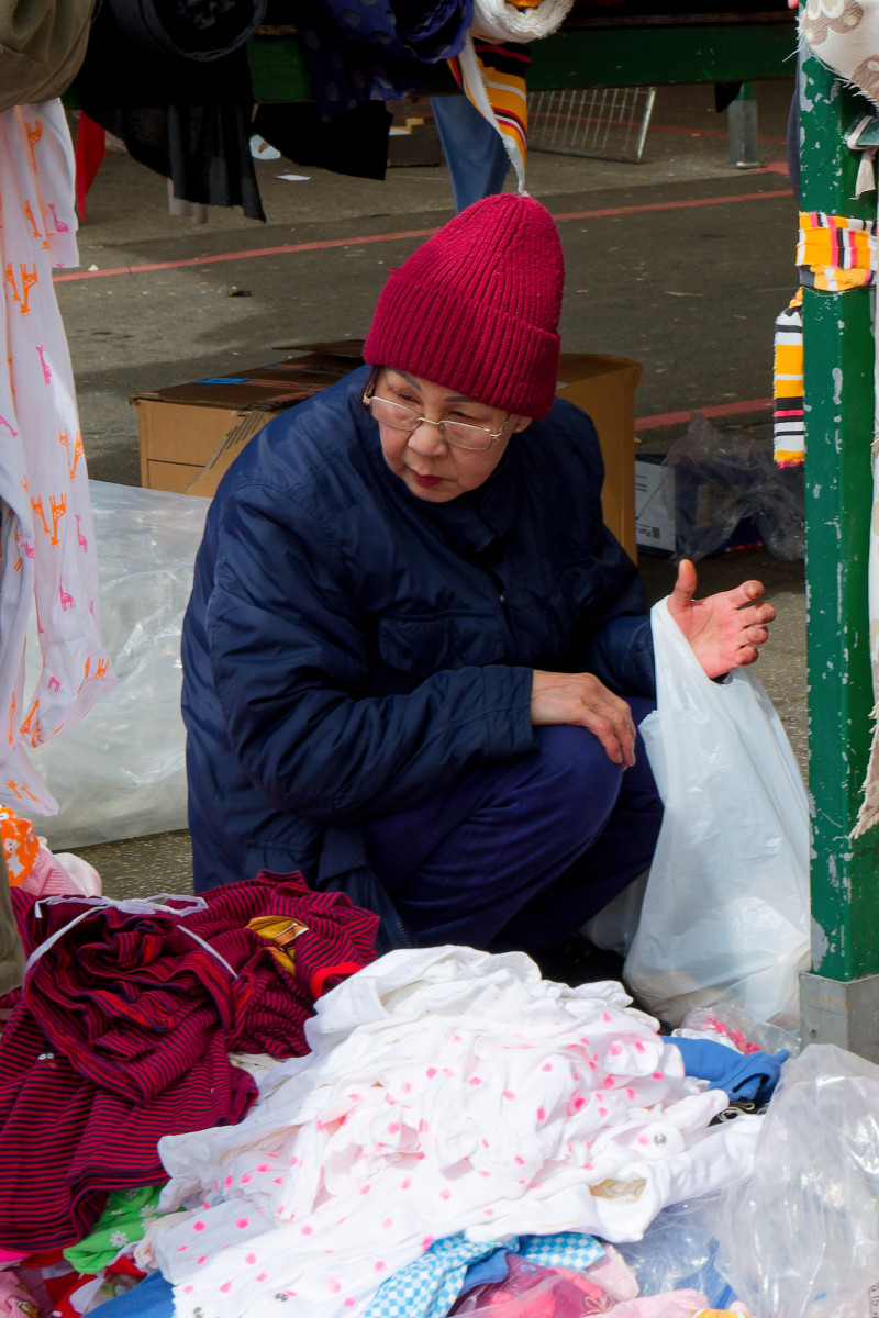 An asian woman dressed with a navy winter coat and read wooly hat is crouching next to a pile of clothes displayed on the ground at the Bullring Market in Birmingham