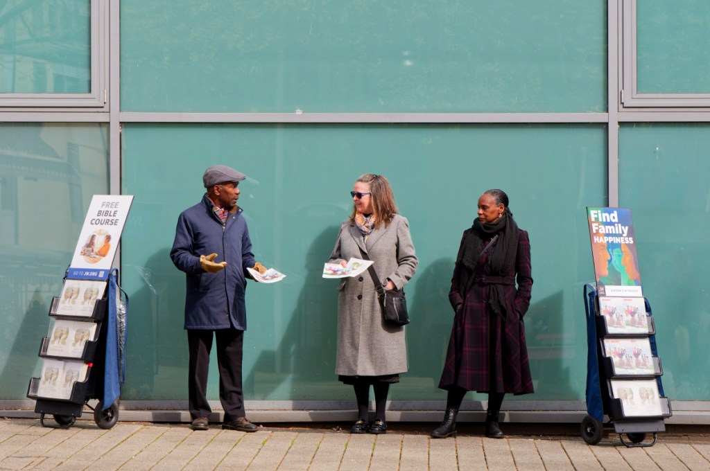 Three Jehovah witnesses, a black man, a black and a white women are standing outside a glass building near the Bullring market in Birmingham. They are wearing winter coats and are displaying literature on both sides.