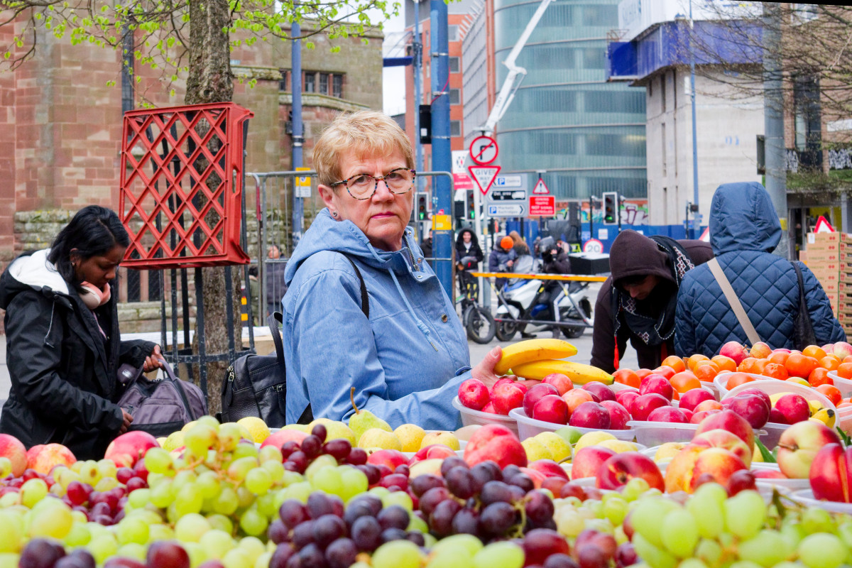 A woman with a light blue jacket is looking at the photographer. She is standing in front of a market stall of coloured fruits. Passersby are walking around.