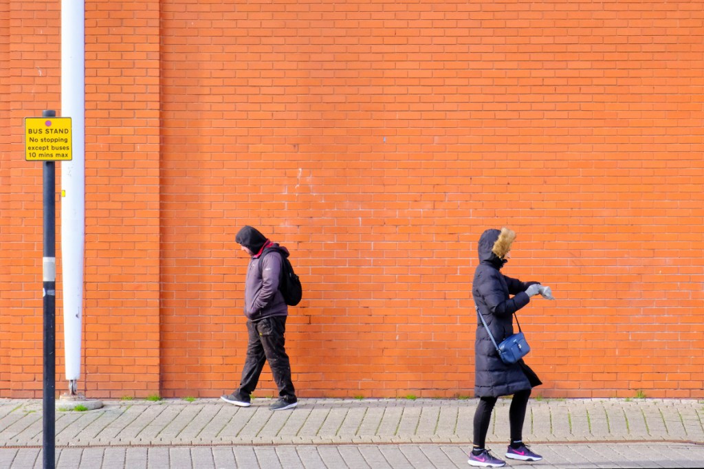 Two passersby are walking in opposite directions in front of a bright red brick wall outside the Bullring Market, Birmingham. They are wearing warm clothes. One is a lady and she is checking her watch.