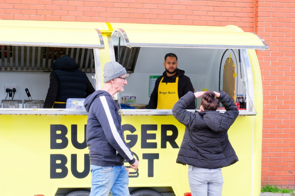 The image shows a yellow Burger fast food wan with a trader with a yellow apron talking to a female customer, while a man is walking in front of.