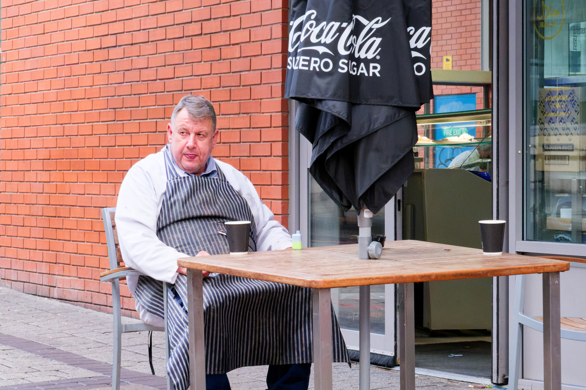 A butcher is having a rest with a drink, seating at a table outside the Bullring Market in Birmingham. He is wearing a striped apron.