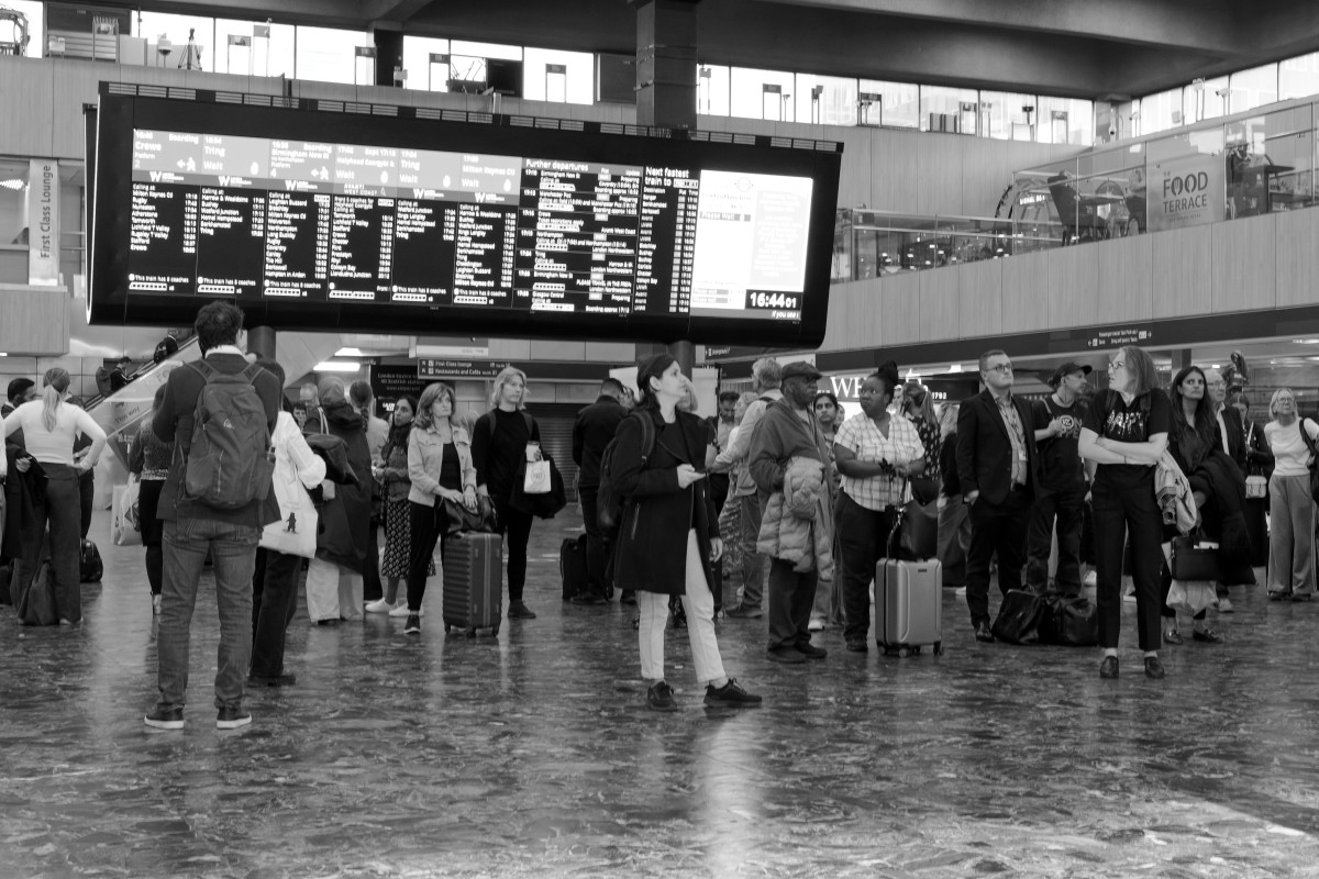 Commuters are waiting for their trains on the Euston station concourse, under the train information screen.