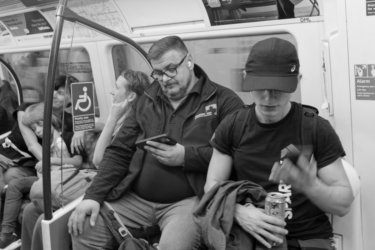 A woman with a cap and a man are commuting on the Bakerloo Line, in London. They are absorbed watching their phones. In the background, a mum is holding a young boy sleeping