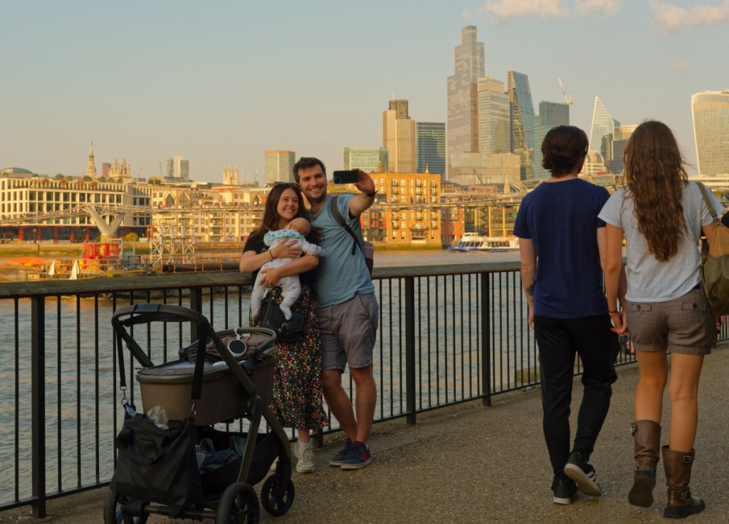 A family of tourists, with a baby, are taking a selfie on the Thames river Southbank, with two tourists walking by and the City in the background.