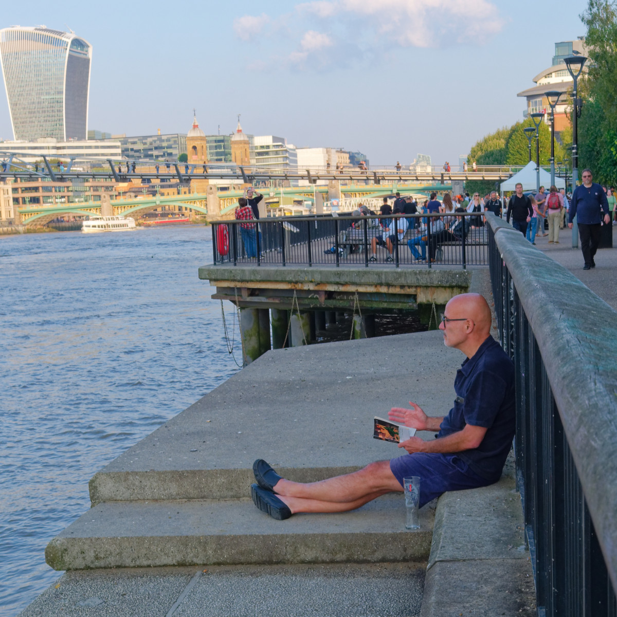 A gentleman in shorts is seated on the bank of the Thames. He is having a book in his left hand and looking in front of him across the river, with the Millennium bridge and the Walkie-talkie building in the background.