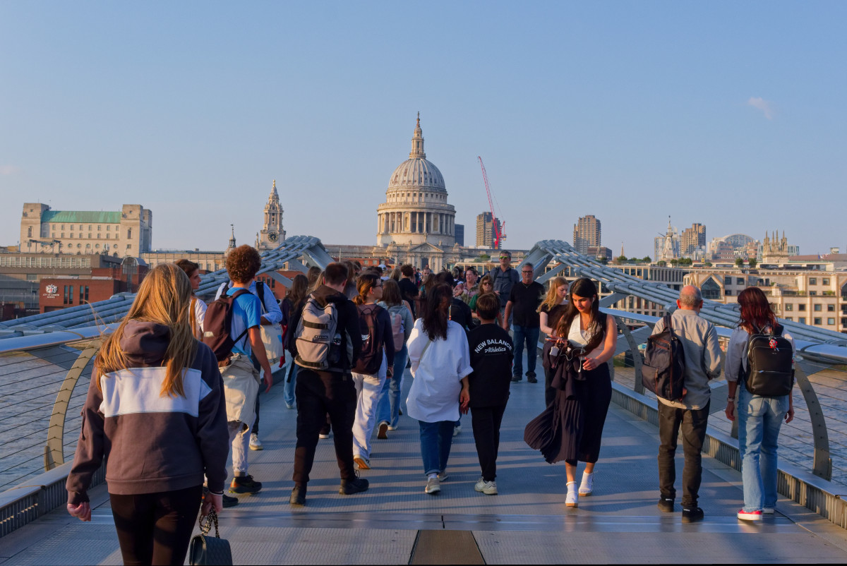 A group of tourists are crossing the Millennium bridge, on a late sunny afternoon, with St Paul's cathedral dome in the background.