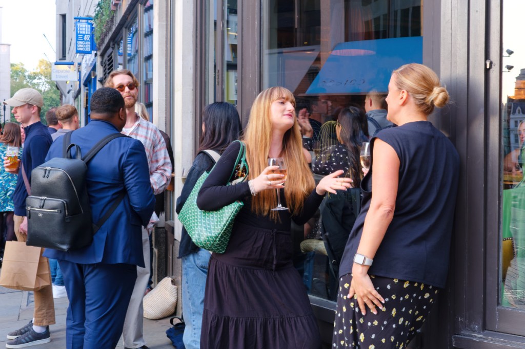 A group of people drinking and chatting outside a bar. A your lady is talking with a friend, with a glass of wine in hand.