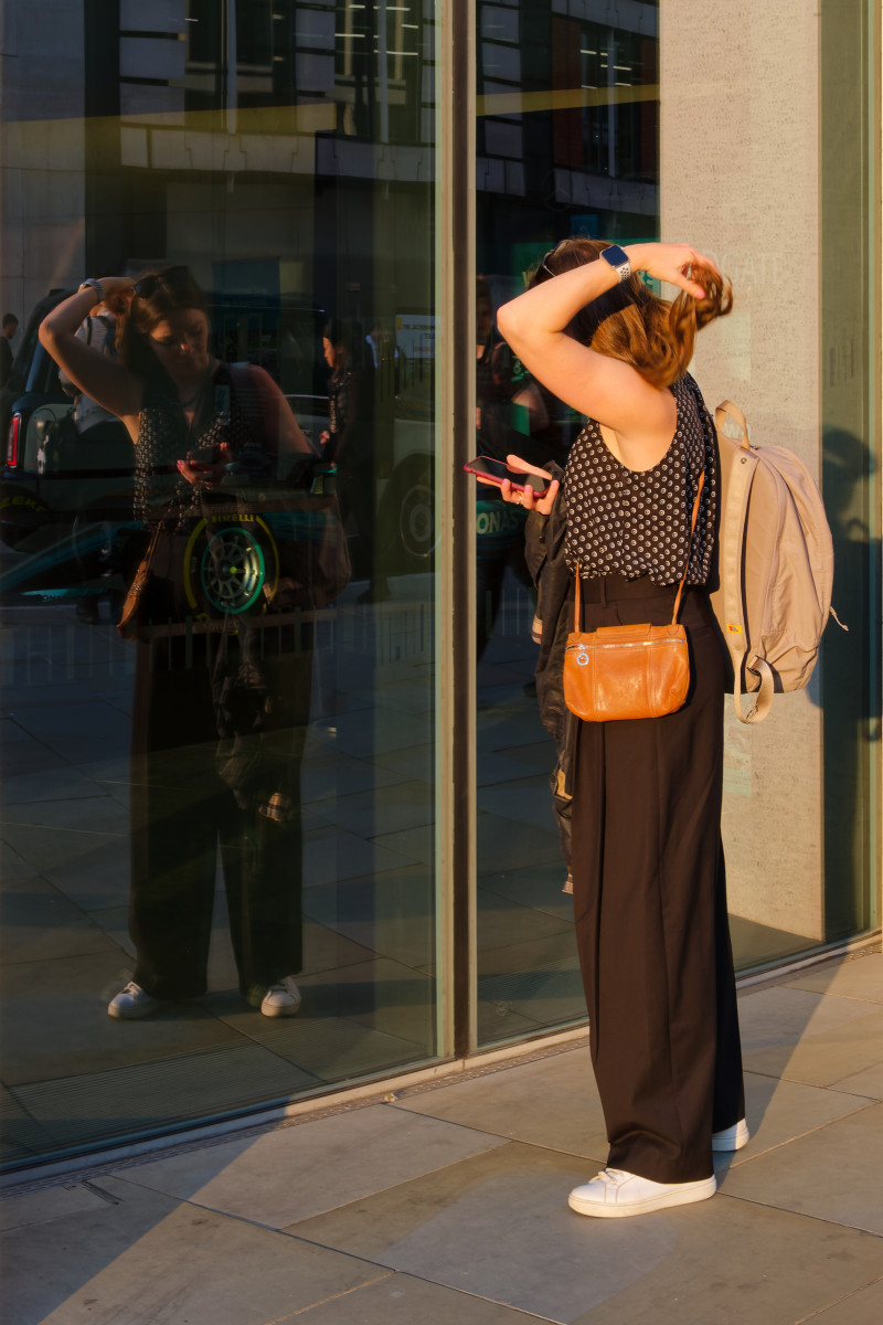 A young woman is looking at her phone and touching her hair, with her reflection in the glass window. The picture was taken mid-September around 5pm with a warm light.