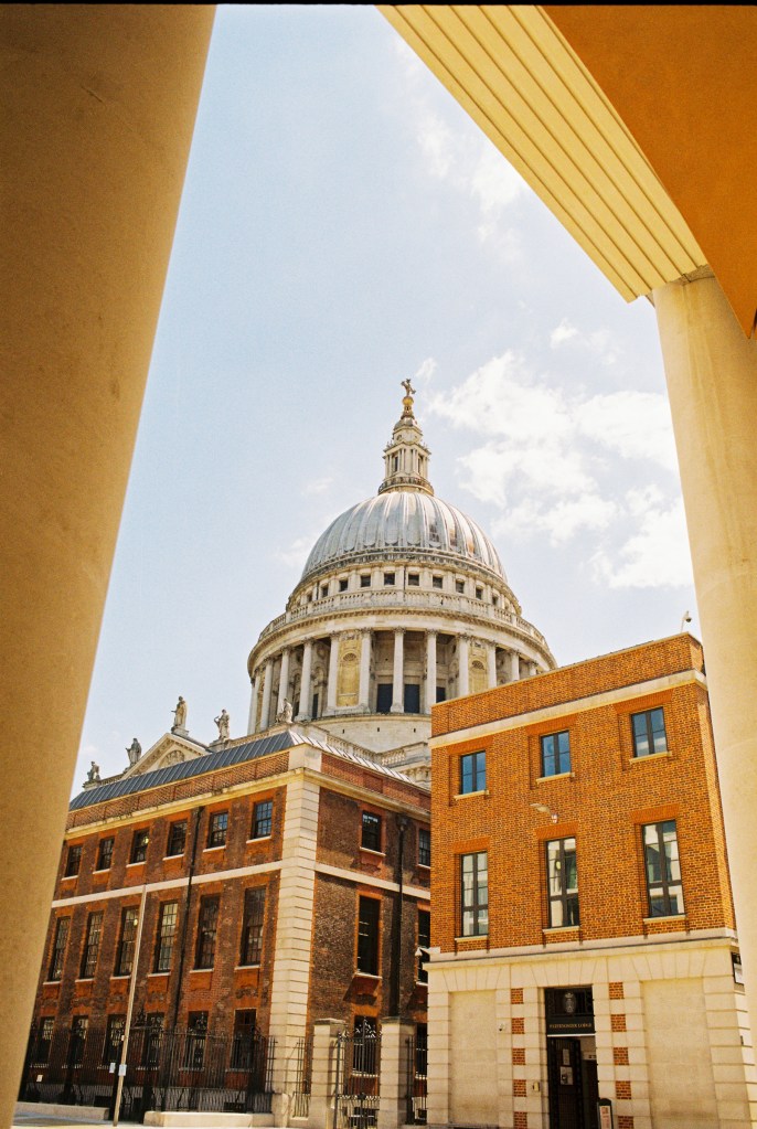 The dome of St Paul's cathedral is shown over modern brick buildings on Paternoster's square. The image is framed by the structure of a building on the square