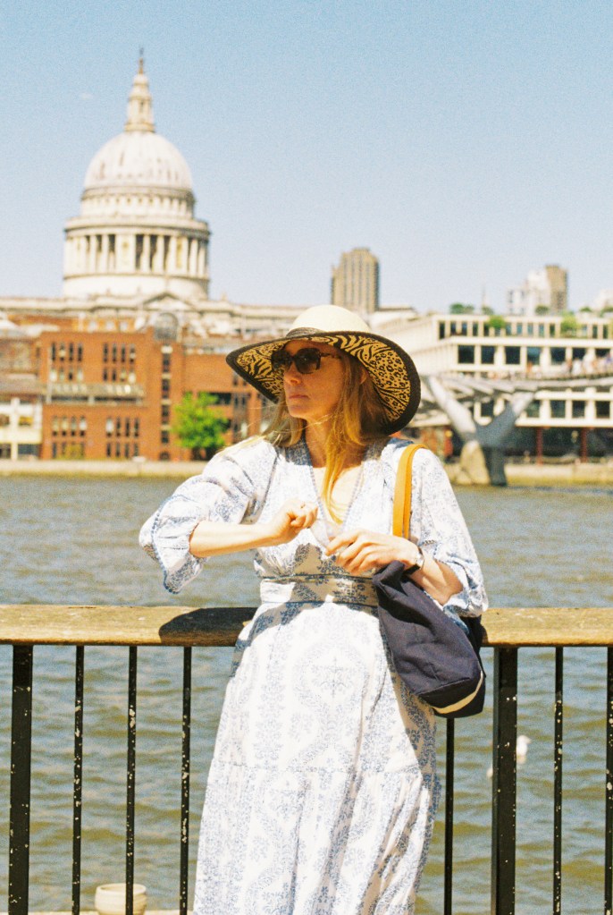 A woman wearing a dress and large hat is standing next to the railing on the Thames pathway with St Paul's cathedral dome in the background