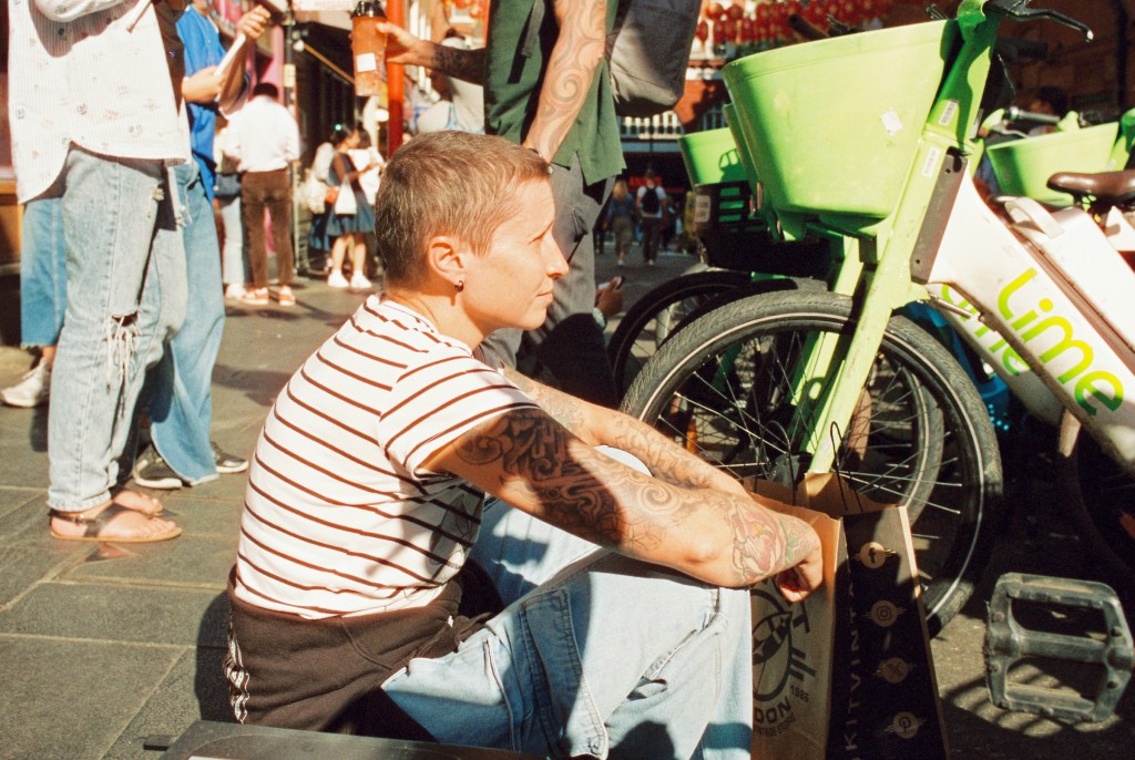 A woman is seated on the pavement edge surrounded by electric bikes. She is wearing a stripe tee-shirt and has tattoos on her arm