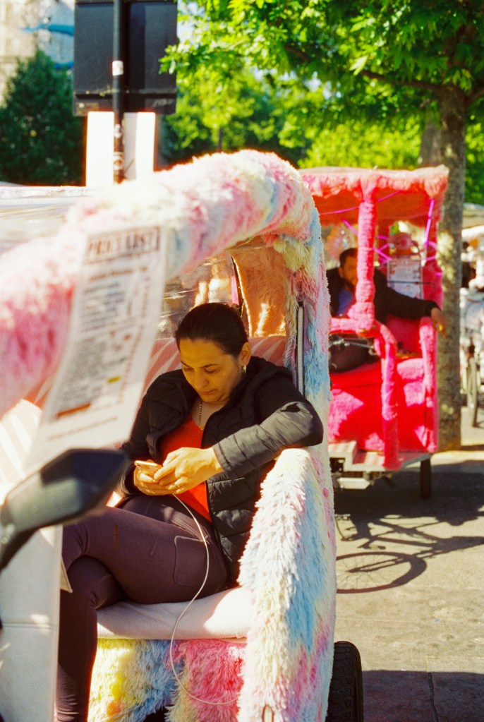 A woman tuk-tuk driver is seated at the back of her vehicle, on her phone, waiting for tourists