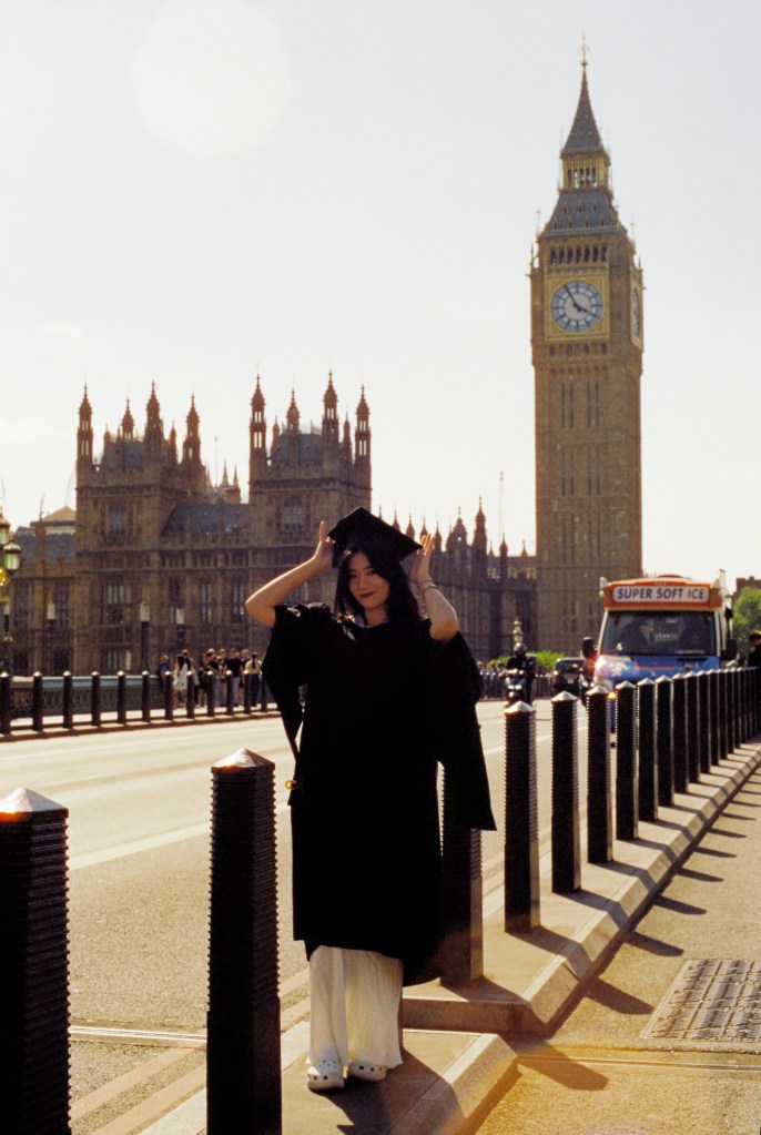 A woman wearing a gown is celebrating her graduation on Westminster Bridge posing in front of Westminster palace.