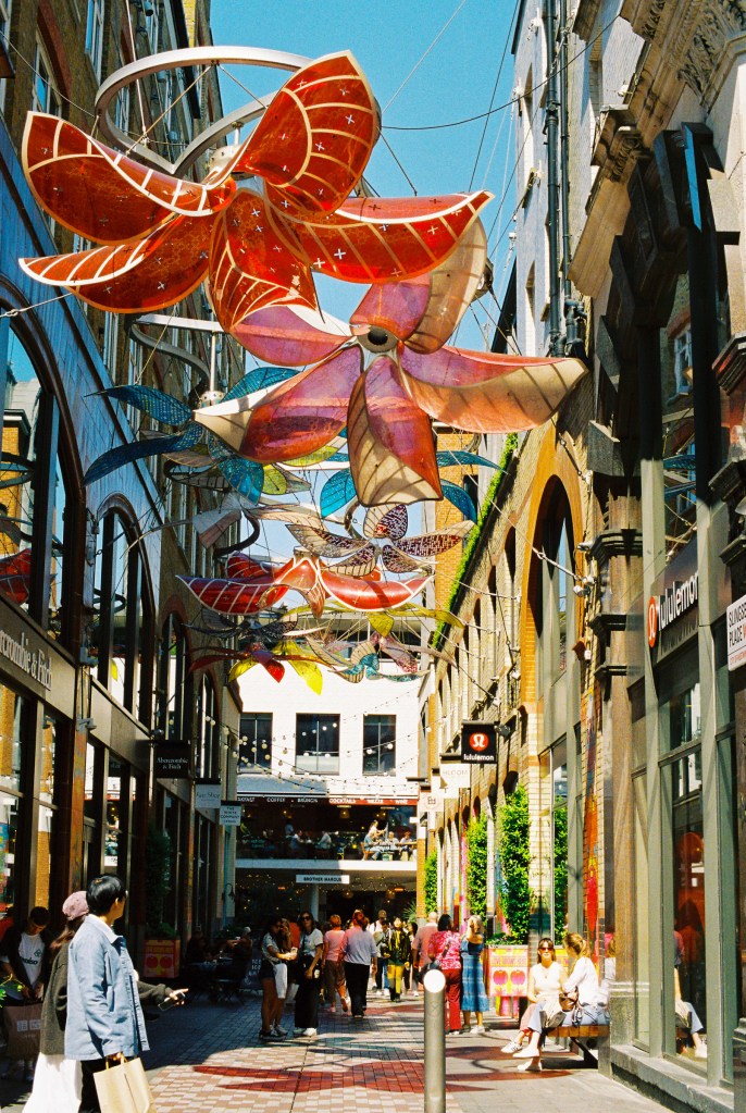 Slingsby Place displays a sculpture of flowers hanging above the passage way with trendy shops on both side.