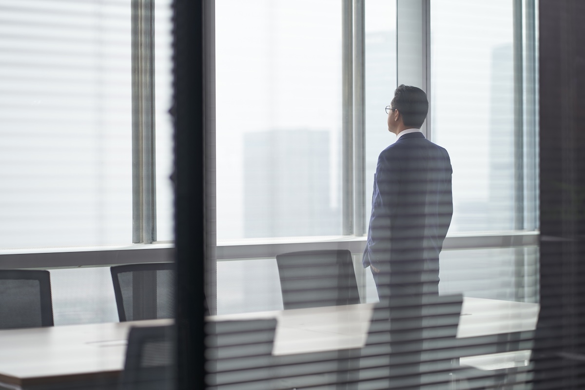 Rear side view of a business man standing in a meeting room in front of office window looking out and thinking hands in pockets
