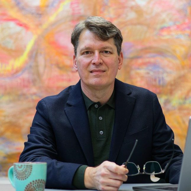 Bruno Vinel seated at a deck with a laptop and a mug, and a colourful background